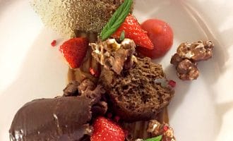 A plated dessert featuring chocolate sponge, chocolate mousse, fresh strawberries, candied nuts, a sugar shard, caramel sauce, and a mint sprig, all artfully arranged on a white plate.