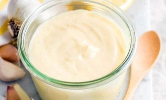 A small glass jar filled with creamy aioli sits on a marble surface, surrounded by garlic cloves, a wooden spoon, a halved lemon, and a white cloth with yellow stripes.