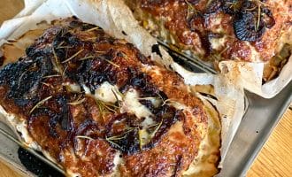 Two baked meatloaves with caramelised, browned tops, garnished with herbs and sliced onions, resting on parchment-lined baking trays. The meatloaves are golden with visible seasoning and a rustic appearance.