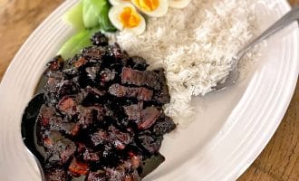 A white oval platter with steamed white rice, soy-braised pork belly in dark sauce, sliced soft-boiled eggs, and blanched green vegetables, served with a large serving spoon.