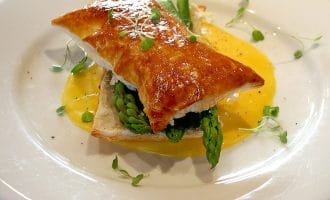 A golden puff pastry sits atop fresh asparagus spears and sauce on a white plate, garnished with microgreens, on a light wooden table. A fork and knife are in the background.