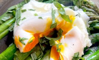 A poached egg with a runny yolk sits atop bright green asparagus spears, garnished with chopped fresh herbs, on a dark plate.