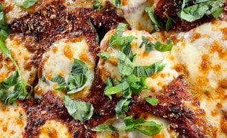 Close-up of baked chicken parmigiana topped with melted mozzarella cheese, tomato sauce, grated parmesan, and fresh basil leaves. The dish has a golden, bubbly, and slightly browned appearance.