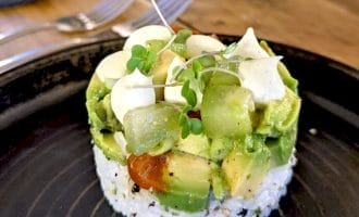 A round stack of sushi rice topped with diced avocado, kiwi, cherry tomatoes, microgreens, and a drizzle of creamy sauce, served on a black plate with a fork and knife in the background.