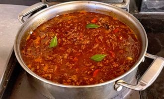 A large stainless steel saucepan on a hob contains simmering meat sauce with visible minced beef, tomato pieces, and bay leaves floating on the surface.
