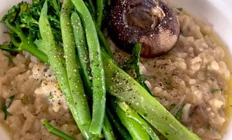 A bowl of creamy risotto topped with steamed tenderstem broccoli and a roasted mushroom, garnished with cracked black pepper and fresh herbs.