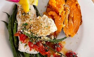 A hand squeezes a lemon over a plate with baked fish topped with breadcrumbs and cherry tomatoes, roasted green beans, and a split baked sweet potato sprinkled with seasoning.