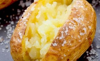 A baked potato split open to reveal its fluffy interior, with coarse salt sprinkled on its crispy golden skin and on the surrounding dark surface.