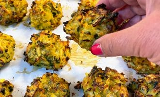 A hand with pink nail varnish picks up a golden-brown baked vegetable fritter from a tray lined with baking paper, surrounded by several similar fritters.