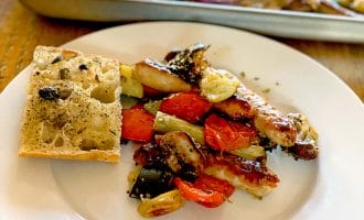 A white plate with roasted vegetables, sausage, and a slice of olive focaccia bread. A baking tray with more roasted vegetables and sausages is in the background on a wooden table.