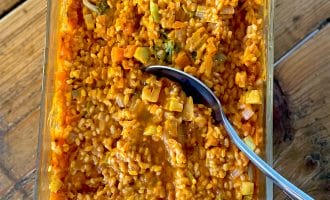 A glass dish filled with creamy orange risotto containing visible pieces of vegetables, such as carrots and broccoli, with a silver spoon resting inside. The dish is placed on a wooden surface.