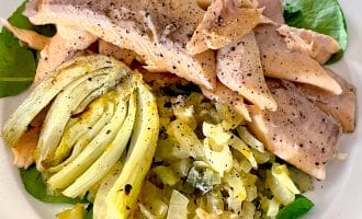 A plate of poached fish fillets topped with black pepper, served on a bed of sautéed greens, onions, and a wedge of cooked artichoke. The dish is arranged on a white plate.