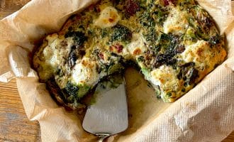 A baked vegetable and cheese frittata in a parchment-lined dish, with a serving spatula and a wedge already removed, sits on a wooden table.