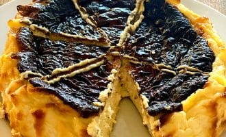 A round, rustic cheesecake with a deeply browned, cracked top sits on a white plate. One large wedge is missing, revealing the creamy interior. The dessert is on a wooden surface.