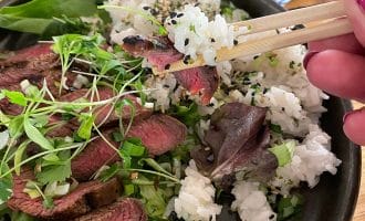 A hand uses chopsticks to pick up a bite of sliced steak and rice from a bowl filled with white rice, sesame seeds, sliced steak, fresh greens, and microgreens.