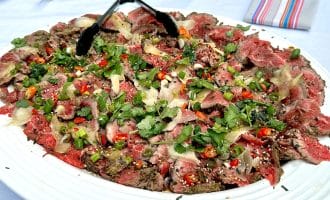 A large white platter of thinly sliced beef carpaccio topped with herbs, red chillies, sesame seeds, and shaved parmesan, with tongs resting on top; a white tablecloth and colourful serviette in the background.