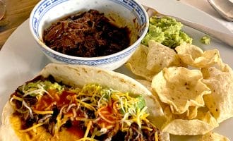A plate with a soft taco filled with shredded beef, cheese, and sauce, alongside tortilla chips, guacamole, and a bowl of shredded beef. A hot sauce bottle is in the background.