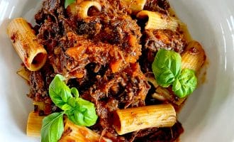 A white bowl filled with rigatoni pasta topped with a rich, chunky meat sauce, garnished with fresh basil leaves.