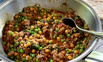 A metal pan filled with a colourful dish of minced meat, green peas, and chickpeas in a tomato-based sauce sits on a wooden table, with a spoon inside. Plates of rice and flatbread are visible in the background.