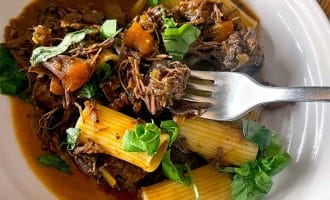 A white plate with rigatoni pasta topped with shredded beef in a rich sauce, diced vegetables, and fresh basil leaves. A fork rests on the plate.