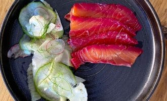 A black plate with slices of beet-cured salmon and a side salad of thinly sliced cucumber, radish, cauliflower, and fresh dill, set on a wooden table.