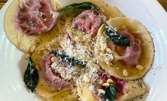A white plate of round ravioli with purple filling, topped with grated cheese, crispy sage leaves, toasted pine nuts, and black pepper, on a wooden table.