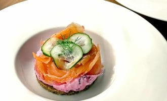 An elegantly plated starter featuring a round base of dark bread topped with a pink creamy spread, slices of smoked salmon, thin cucumber slices, and a sprig of dill, served on a white plate.