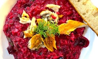 A white bowl of vibrant beetroot risotto, topped with chunks of smoked fish and fresh dill, served with a slice of toasted bread on the side.