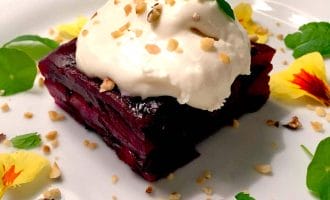 A square slice of beetroot terrine topped with a dollop of cream and chopped nuts, garnished with yellow edible flowers and fresh mint leaves on a white plate.