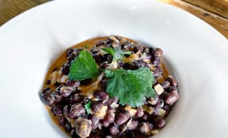 A white bowl filled with creamy black beans garnished with fresh coriander leaves, placed on a wooden table.