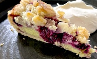 A slice of berry crumble pie with a crumbly topping sits on a dark plate next to a dollop of whipped cream.