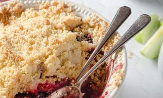 A round baking dish filled with berry crumble, topped with a golden, crumbly crust. Two spoons rest inside, and some fruit filling is visible. In the background, a bowl of berries and apple slices are partially seen.