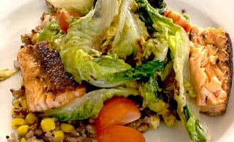 A plate of grilled salmon served with mixed grains, sweetcorn, cherry tomatoes, and sautéed greens, arranged on a white dish.