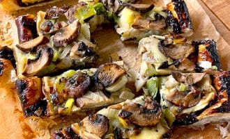 Rectangular slices of savoury tart topped with mushrooms, leeks, and cheese on a sheet of baking parchment, resting on a wooden surface. The tart has a golden, flaky pastry and appears freshly baked.