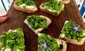 Sliced bread topped with creamy cheese, chopped green peas, herbs, and purple edible flowers arranged on a wooden board, with books visible in the background.