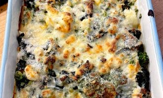 A baked casserole dish filled with broccoli, cauliflower, and melted cheese, browned on top, sitting on a wooden surface.