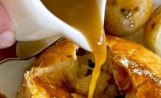 Golden brown gravy is being poured from a small jug over a flaky pastry dish, with roast potatoes visible in the background. The pastry is cracked open, revealing its filling inside.