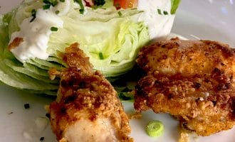 A plate with a wedge of iceberg lettuce topped with creamy dressing, chopped chives, and tomato, served alongside two pieces of crispy fried chicken.