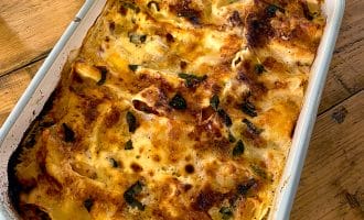 A baked dish of golden-brown lasagne with melted cheese and herbs sits in a white rectangular baking dish on a wooden table.