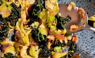 Sautéed cabbage and kale with onions and garlic in a blue speckled bowl, partially eaten, with a metal spoon resting on the side.