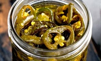A close-up of a glass jar filled with sliced pickled jalapeños in brine, showing seeds and pepper rings. The jar sits on a dark wooden surface.