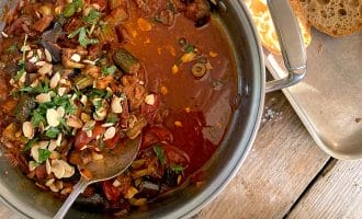 A pot of vegetable stew topped with fresh herbs and sliced almonds sits on a wooden table next to a tray of sliced bread and a plate with green beans.