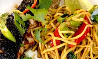 Grilled fish fillet topped with herbs, served alongside stir-fried noodles with red peppers, spring onions, and coriander, garnished with a lime wedge on a white plate.