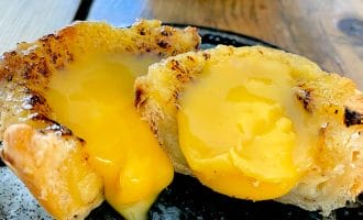 A close-up of a pastry cut in half, revealing a rich, yellow, creamy filling oozing out onto a dark ceramic plate. The pastry has a golden, flaky crust and sits on a wooden table.
