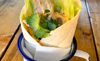 A tortilla wrap filled with lettuce, chicken goujons, creamy sauce, and fresh coriander, served upright in a white enamel mug on a wooden table.