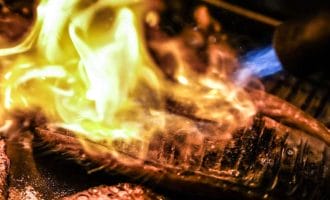 Slices of fish being seared with an open flame, creating a dramatic burst of yellow and orange fire over the surface, highlighting the texture and cooking process in a close-up shot.