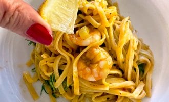 A hand with red nail varnish squeezes a lemon wedge over a plate of prawn pasta with yellow sauce, garnished with fresh herbs.
