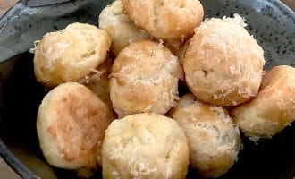 A dark bowl filled with round, golden-brown cheese puffs topped with grated cheese, sitting on a wooden surface.