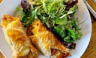 A white plate with two golden, flaky pastry rolls filled with cheese and meat, served alongside a fresh mixed green salad, set on a wooden table with a fork and knife.