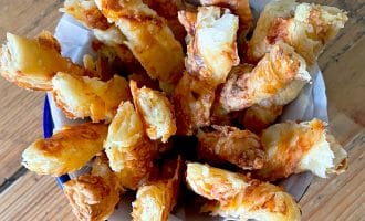 A bowl lined with greaseproof paper filled with golden, flaky cheese straws made from puff pastry, arranged upright on a wooden table.